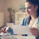Smiley young girl at home holding document and typing on laptop. Close up.