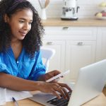 Candid shot of focused beautiful young African housewife wearing headband and casual shirt paying utility bills online using laptop computer, sitting at kitchen table with papers and smiling